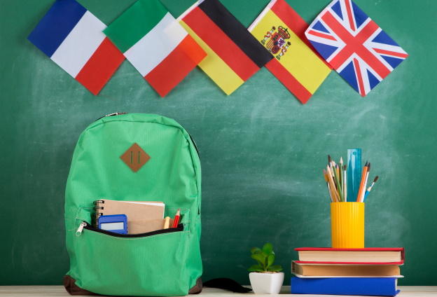 Güner Rucksack auf Tisch mit Büchern und Schreibzubehör und den Flaggen von Großbritannien, Spanien, Deutschland, Italien und Frankreich im Hintergrund.