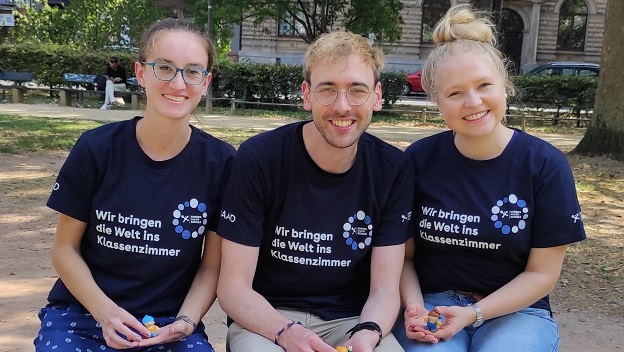 3 junge Personen mit einem EmS-Shirt sitzen in einem Park auf der Bank und lächeln in die Kamera