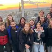 Junge engagierte Studierende stehen auf dem Triangel Turm in Köln und lächeln in die Kamera. Im Hintergrund sieht man den Sonnenuntergang überm Dom.