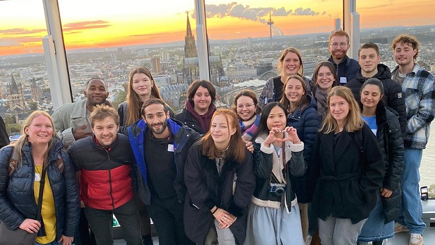 Junge engagierte Studierende stehen auf dem Triangel Turm in Köln und lächeln in die Kamera. Im Hintergrund sieht man den Sonnenuntergang überm Dom. 