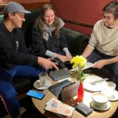 3 junge Studierende sitzen in einem Café an einem Tisch mit einem Tablet, Notizbüchern und Europa macht Schule Flyern und besprechen alles für das kommende EmS-Programmjahr in Jena