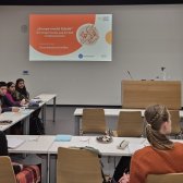 Man schaut von hinten auf junge Studierende an Tischen in U-Form sitzen. Vor ihnen wird eine PowerPoint vom Programm 