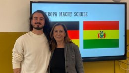 Internationaler Student und Lehrerin stehen vor einem Bildschirm, der die deutsche und die bolivianische Flagge anzeigt.